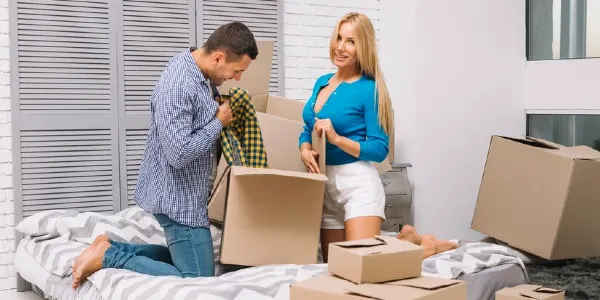 A couple is packing cloths in a moving box on a bed inside a bedroom