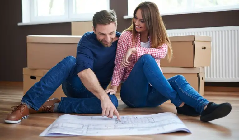 A couple on floor pointing towards a floor plan paper with moving boxes.